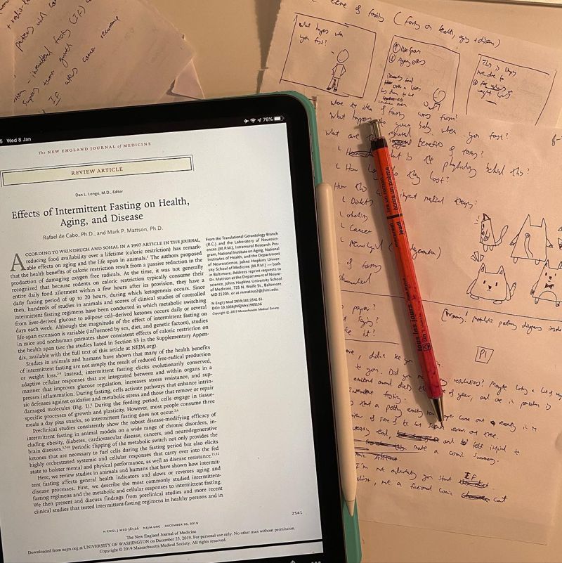 Desk with NEJM fasting paper on iPad alongside Professor Cat comic sketches